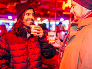 Two people in winter clothing holding cups of Brothers Mulled Festive Cider with a colourful, blurred background