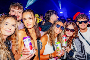 Group of friends at a festival holding cans of Brothers Cider with colourful lights in the background