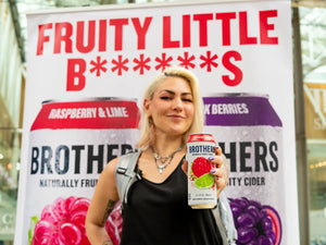 Person holding a Raspberry & Lime Brothers cider can in front of a branded backdrop Fruity Little