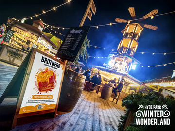 Outdoor winter market scene with festive lights and a sign for 'Brothers Spiced Toffee Apple Cider' at Hyde Park Winter Wonderland.
