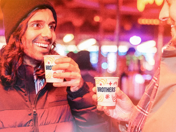 Two people smiling and holding paper cups with the 'BROTHERS' logo, set against a warm, blurred night winter market background with colourful lights.