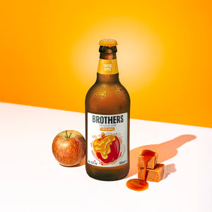 Brothers Toffee Apple cider bottle with an apple, toffee pieces, and caramel on a white surface with orange background