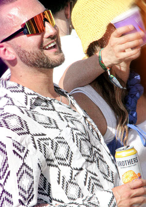 Man wearing patterned shirt and colorful sunglasses holding a can, with a woman in the background.