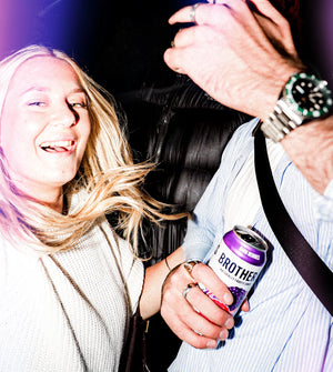 Two people holding a can of Brothers Dark Berries Cider with a dark background at a party